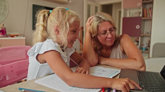 Preschool child studies at a desk with her mother and a laptop. Modern home education and early school preparation