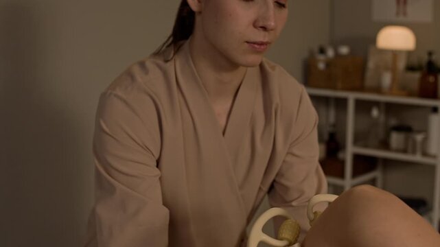 Tilt up closeup of female therapist performing leg massage with wooden tool on female client lying on treatment bed in cozy clinic room