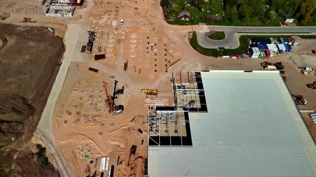 Aerial reveal over large industrial factory construction site showing steel frame erection, orange drilling rig, cranes, foundation piles and staged building materials