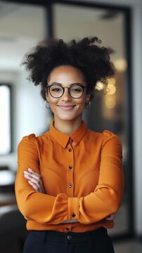 A woman with curly hair and glasses is smiling. She is wearing an orange shirt and has her hands on her hips