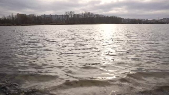 Close up of gentle waves lapping on a muddy shore with dry leaves. Calm lake water ripples with a soft sun reflection. Peaceful, tranquil, serene nature background video for relaxation meditation