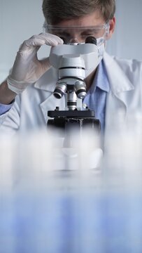 Man scientist wearing lab coat, white gloves, medical mask and protective glasses using microscope doing research in modern laboratory, portrait view. Low key lighting photo. Science and Medicine
