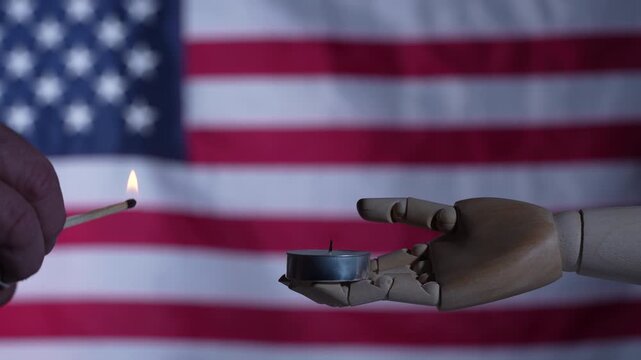 Symbolic image of a wooden mannequin hand holding a burning candle against blurred USA flag. Concept for national mourning memorial day prayer and patriotism