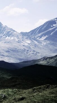 The view shows tall mountains with snow covering their peaks. A green valley spreads out below with rocks and grass in the foreground. Bright sunlight illuminates the scene.