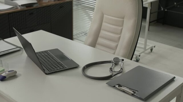No people shot of white office desk with open laptop, stethoscope, grey file folder and non-contact thermometer next to leather chair in clinical setting