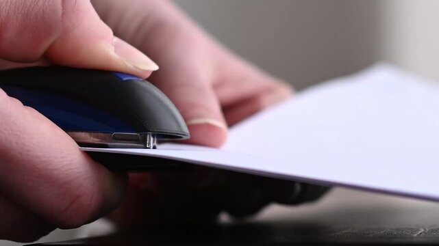 young caucasian woman in warm plaid shirt holding in hands blue stapler and stapling sheets of paper, selective focus, close-up view of hands