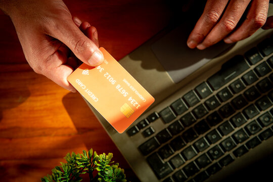 High angle close up of male hands holding an orange credit card while using a laptop keyboard for digital transaction and online shopping.
