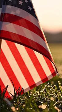 American flag planted in dewy grass at dawn, faintest flag movement, pale amber-pink dawn sky as generous copy space above, Memorial Day solemn and dignified morning atmosphere.