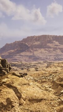 Two hikers walk on rocky ground in a vast desert area. A large mountain stands tall in the background. The sun shines bright above, illuminating the scenery.