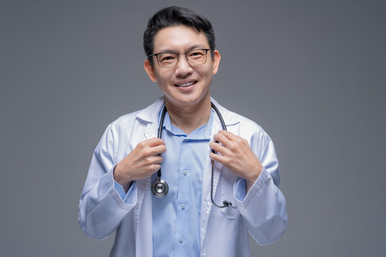 Asian doctor in a white coat with a stethoscope around his neck and glasses, in front of a gray background, smiling at the camera and holding his lapels with both hands.