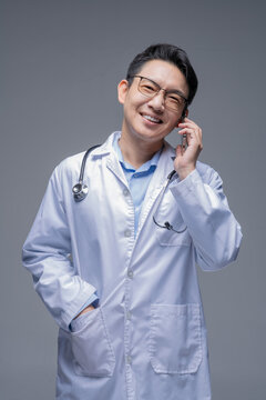 Asian doctor in a white coat with a stethoscope and glasses, isolated on a grey background, talking on a smartphone with a smiling expression and one hand in his pocket.