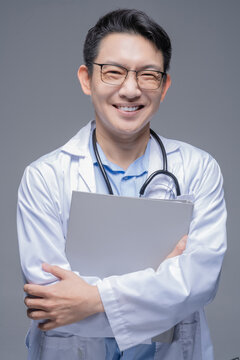 Asian doctor in a white coat with a stethoscope around his neck and glasses, in front of a gray background, smiling at the camera and holding his lapels with both hands.