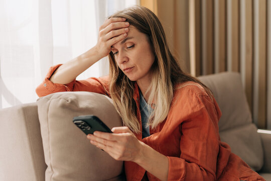 Upset anxious woman sitting on the sofa and looking at her mobile phone.