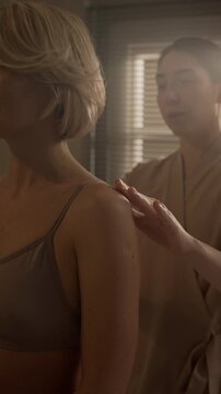 Vertical waist up shot of female orthopedic specialist standing behind Caucasian blonde woman while evaluating her back muscles and posture during consultation in cozy treatment room