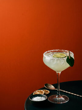 Chilled margarita cocktail with salt rim and lime garnish presented on a dark surface with dried lime and salt dish against a vibrant orange background creating an eye catching visual