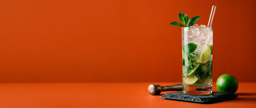 Refreshing mojito cocktail with fresh mint leaves and crushed ice served in a tall clear glass on a slate coaster with a lime wedge and muddler on an orange studio background for summer