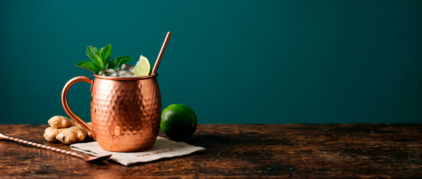 Moscow mule cocktail served in a hammered copper mug adorned with fresh lime and mint garnishes rests on a rustic wooden table alongside ginger and a spoon illuminated by studio lighting