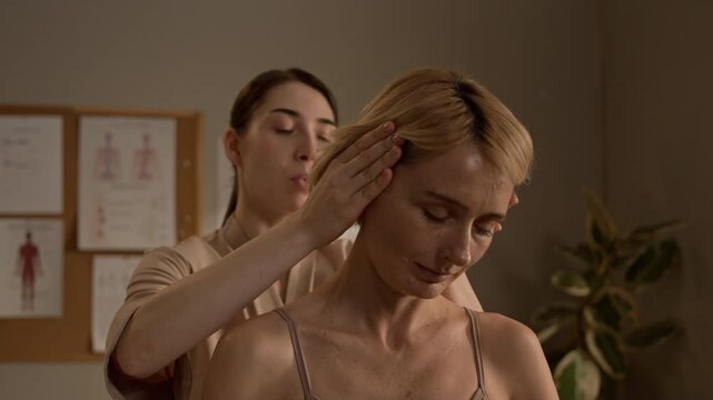 Medium closeup of female massage specialist examining and adjusting female client neck posture in warm wellness interior before therapeutic massage