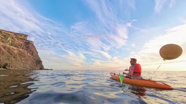 Kayaking ocean sunset man in an orange kayak paddles across calm sea waters alongside a dramatic cliff under a beautiful evening sky