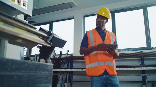 Metal industry worker in a yellow hard hat, monitoring automated system operation, making adjustments via tablet to optimise processes.