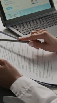 Vertical close-up shot of hands of anonymous physician carefully studying medical service agreement at white table with laptop in background