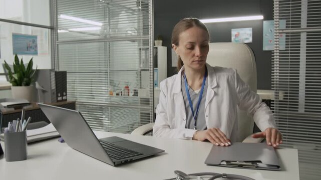Caucasian female gynecologist opening file folder to look at medical report while using laptop at white desk during workday in reproductive center