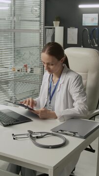 Vertical shot of Caucasian female physician wearing uniform looking at service agreement and using laptop while sitting at white table in leather chair in her office