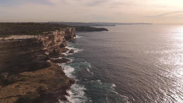 Breathtaking drone footage captures the rugged Malabar Headland cliffs at sunrise as the morning light illuminates the sandstone rocks and waves crash against the Sydney coastline below