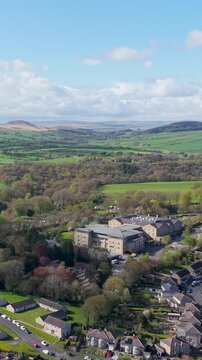 Reversing vertical drone shot pulls back from the Skipton Building Society headquarters, revealing northern Skipton's residential streets, green fields and rolling moorland under blue skies. Spring.