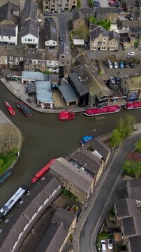 Vertical drone tilts down over Skipton's historic canal basin on Leeds and Liverpool Canal, with vivid red leisure cruise boats, stone buildings, car parks and converging roads. North Yorkshire, spr