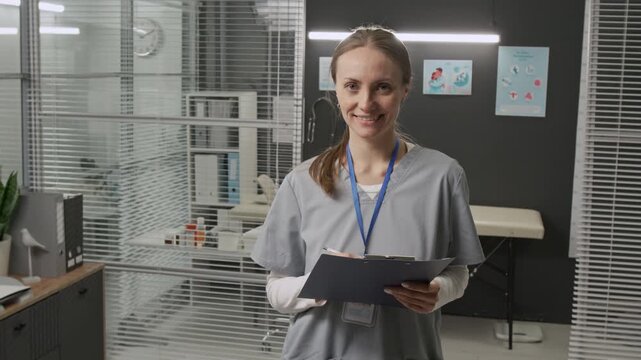 Waist-up portrait of Caucasian female medical specialist wearing scrubs and badge holding clipboard smiling for camera at workplace