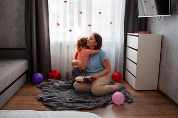 Naklejka premium Wide shot of little Caucasian girl hugs her mother, sits on a carpet in room and reads book. Woman and her daughter spend happy time. Mother's Day Concept