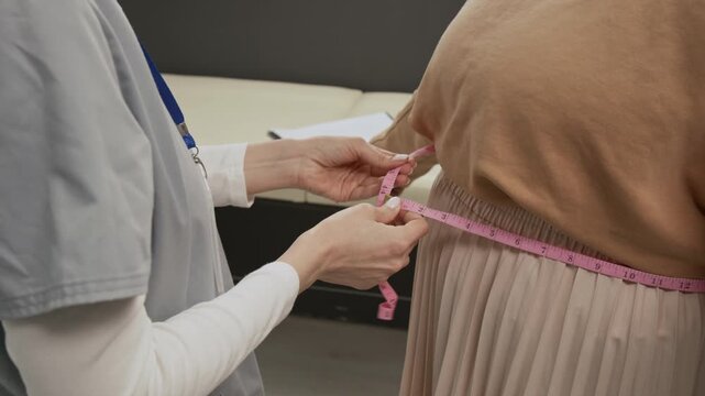 Midsection view of unknown female doctor wearing scrubs using pink sewing tape measuring waist circumference of anonymous patient dressed in sweatshirt and skirt