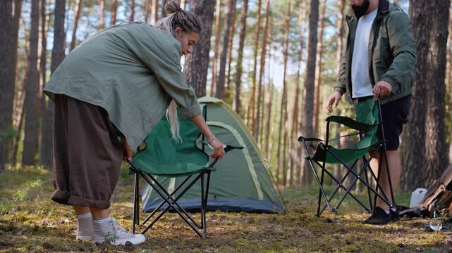 Caucasian man and woman setting up folding chairs and a table near their tent in a sunny pine forest, getting their camp ready for a relaxing outdoor vacation with a guitar nearby