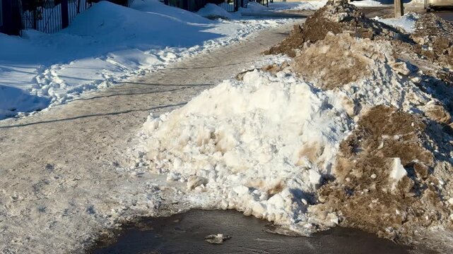 Snow pile beside icy road, sunlit dirty mound from plow at road shoulder, asphalt patch and curb visible, grit and salt residue