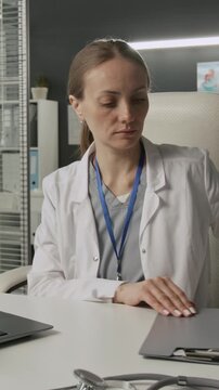 Vertical shot of female medical worker in white coat looking through document with colourful diagram and typing on laptop at workplace