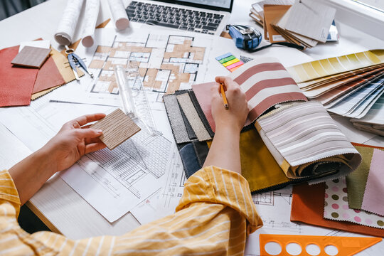 Interior design process with hands selecting wood sample over architectural plans, floor plan, laptop and color palette, residential renovation planning and material selection concept.