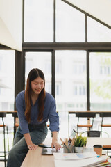 Obraz premium Young Asian businesswoman freelancer smiling while leaning on a desk, using a tablet and laptop in a bright, contemporary office setting, performing online tasks and managing projects