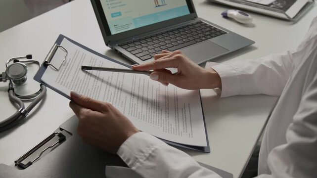Midsection view of unrecognisable female doctor in white coat reading medical contract following lines with pencil while sitting at office desk featuring laptop and stethoscope