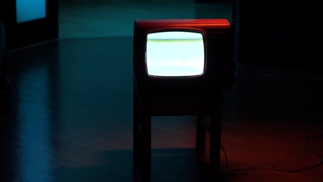 Vintage red television set with a bright glowing cathode ray tube screen sitting on a dark floor with blue and red ambient light creating a mysterious, nostalgic atmosphere