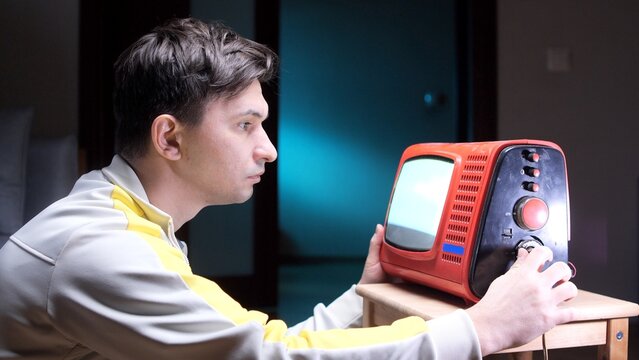 Man in athletic apparel intently watching and adjusting a retro red portable television, experiencing a moment of 80s or 90s nostalgia while controlling the old technology