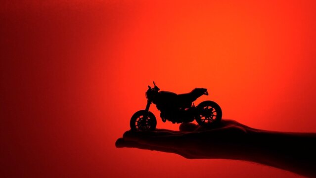 Hand silhouette holding a small toy motorcycle casting a dark shadow against a vibrant red backdrop, representing freedom, hobby, and the dream of adventure