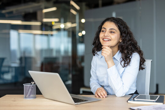 Smiling young Hispanic businesswoman sitting at a desk with a laptop and dreaming about success. Happy diverse female entrepreneur thinking of new ideas in modern office.