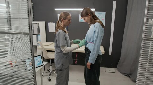 Gynecologist measuring waist circumference of gen Z woman for health assessment in brightly lit medical office setting