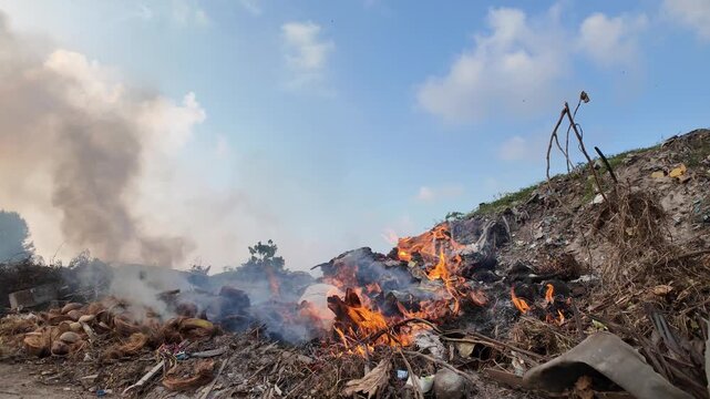 Smoke billows from a burning landfill where waste material ignites under a cloudy sky.