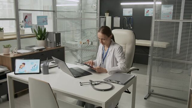 Female doctor in white coat looking through medical form and entering data while working on laptop at office desk featuring stethoscope, file folder and digital tablet in fertility clinic