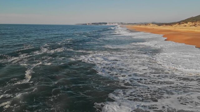 Aerial dolly forward shot of the Atlantic Ocean waves crashing on the sandy coastline. This scenic view features Praia do Salgado beach near Nazar&eacute;, Portugal, on a bright and sunny day