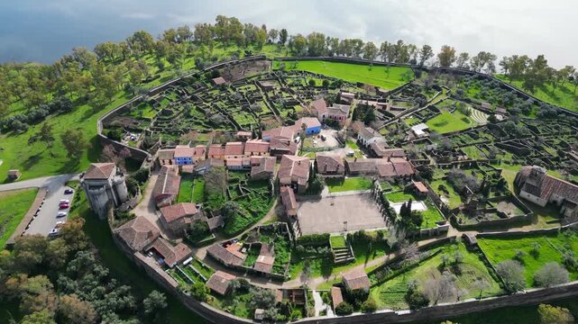 Drone ascending view over Granadilla walled village in Spain, revealing medieval layout, rooftops and fortified walls surrounded by natural landscape.