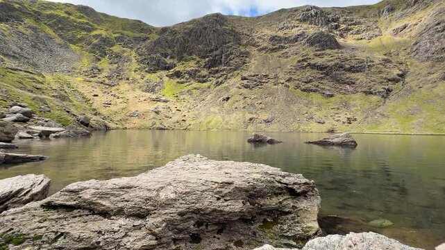 A static ground-level shot of Low Water tarn at Coniston, framed by a large boulder in the foreground and steep rocky crags rising dramatically on all sides. Lake District, Cumbria.