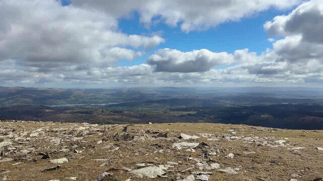A right pan from a rocky Lake District mountain summit reveals a sweeping panorama of fells, distant valleys, and lakes under large white clouds and vivid blue skies. Cumbria, England.